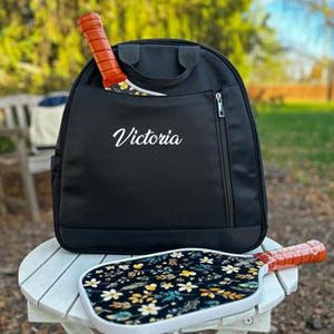 K&ouml;nnte beinhalten: Schwarzer Rucksack mit einer Rei&szlig;verschlusstasche und einem Paddelschl&auml;ger im Inneren. Der Rucksack hat den Namen "Victoria" auf der Vorderseite gestickt.
