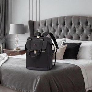 May include: A black backpack with gold-colored zippers and accents. The backpack has a top handle and adjustable shoulder straps. It is sitting on a gray bedspread in a bedroom setting. The bed has a tufted headboard and decorative pillows.