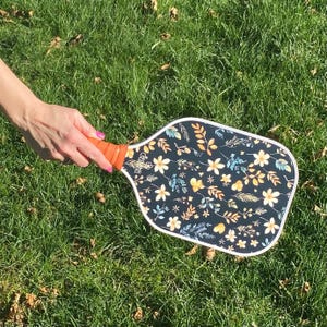 K&ouml;nnte beinhalten: Ein wei&szlig;-schwarzer Schl&auml;ger mit einem floralen Design mit gelben, blauen und orangefarbenen Blumen. Der Schl&auml;ger hat einen orangefarbenen Griff.