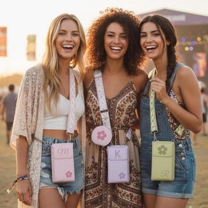 May include: Three smiling people pose outdoors, each holding a crossbody phone bag. The bags are pastel pink, lavender, and green, with a flower design and a letter. The bags have matching straps. The women are wearing casual clothing.