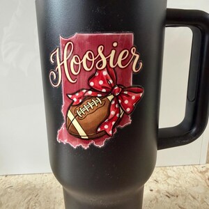 May include: Black travel mug with a handle, featuring a design with the word "Hoosier" in cursive, a football, and a red bow with white polka dots. The design is set against a red outline of the state of Indiana.
