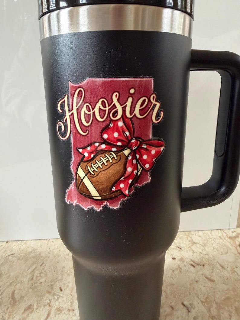 May include: Black travel mug with a handle, featuring a design with the word "Hoosier" in cursive, a football, and a red bow with white polka dots. The design is set against a red outline of the state of Indiana.