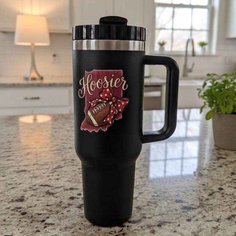 May include: Black travel mug with a handle and a lid. The mug features a graphic of the state of Indiana with the word "Hoosier" and a football. The graphic is red, white, and brown.