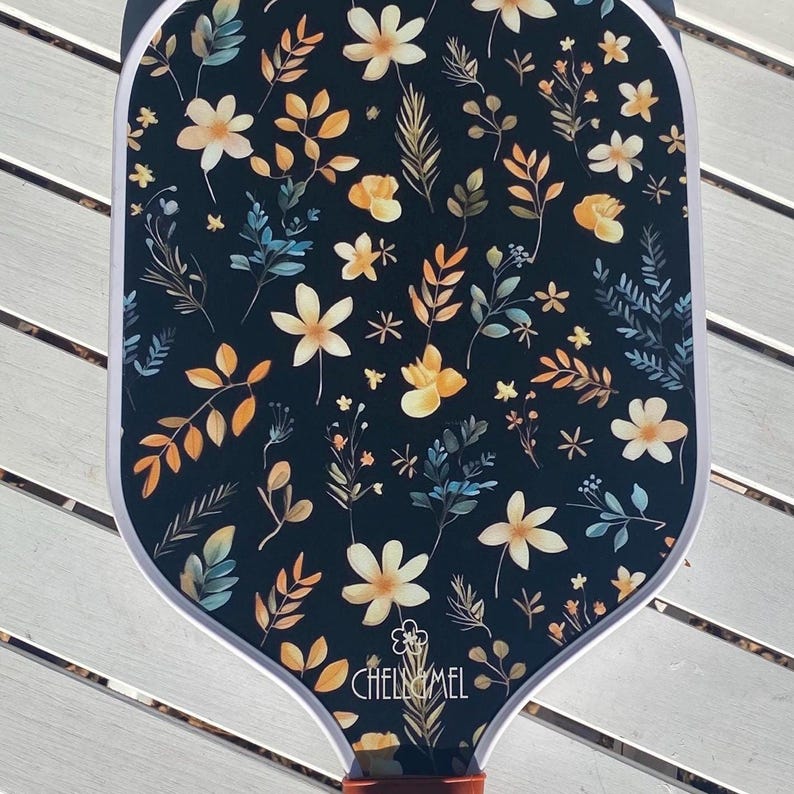 K&ouml;nnte beinhalten: Ein schwarzer Schl&auml;ger mit wei&szlig;em Rand und einem floralen Muster in Gelb-, Orange- und Blaut&ouml;nen. Der Schl&auml;ger hat den Text "CHELLEMEL" unten aufgedruckt.