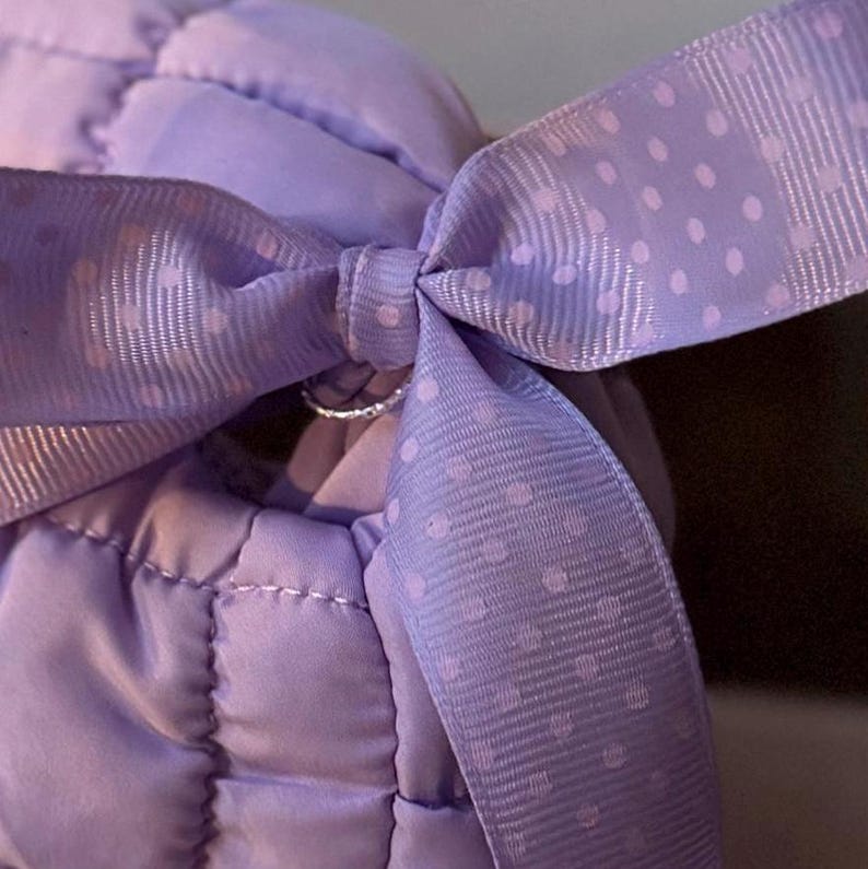 May include: Close-up of a lavender quilted fabric with a matching ribbon tied in a bow. The ribbon features a delicate polka dot pattern. The stitching on the fabric is visible, adding texture to the design. The overall aesthetic is soft and feminine.