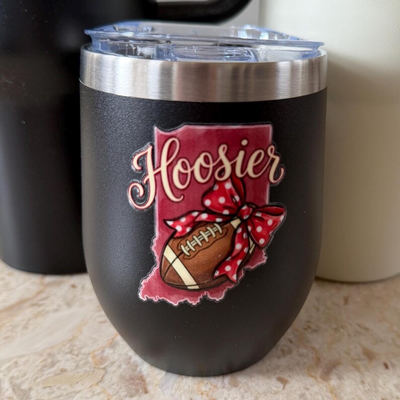 May include: Black insulated tumbler with a silver rim and clear lid. The tumbler features a graphic of the state of Indiana in red, with the word "Hoosier" in cursive and a football with a red polka dot bow.