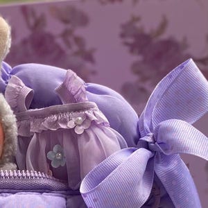 May include: A close-up of a lavender-colored doll dress with a matching bow. The dress features a ruffled bodice, a sheer skirt, and is adorned with a pearl-centered flower. The bow has a polka-dot pattern. The background is a floral design.