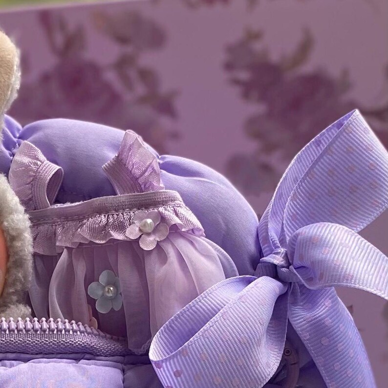 May include: A close-up of a lavender-colored doll dress with a matching bow. The dress features a ruffled bodice, a sheer skirt, and is adorned with a pearl-centered flower. The bow has a polka-dot pattern. The background is a floral design.