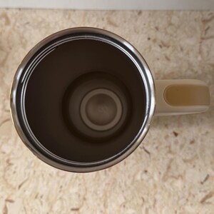 May include: Stainless steel travel mug with a cream-colored handle. The interior of the mug is a dark brown color. The mug is sitting on a light brown surface.