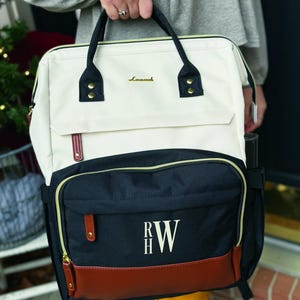 May include: A two-tone backpack featuring a cream-colored upper section and a black lower section with a brown faux leather base. The bag displays the gold text "Lovook" and the embroidered monogram "RW" in white. It includes black handles and zippers.
