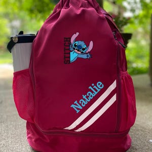 May include: A maroon drawstring backpack featuring a blue cartoon character and the name "Natalie" in light blue. The bag has white stripes and mesh side pockets, with a water bottle in one pocket.