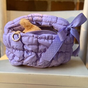 May include: A quilted lavender bag with a zipper closure and a light brown stuffed animal peeking out. The bag has a matching lavender ribbon with white polka dots tied in a bow. A gold-colored ring is attached to the side.