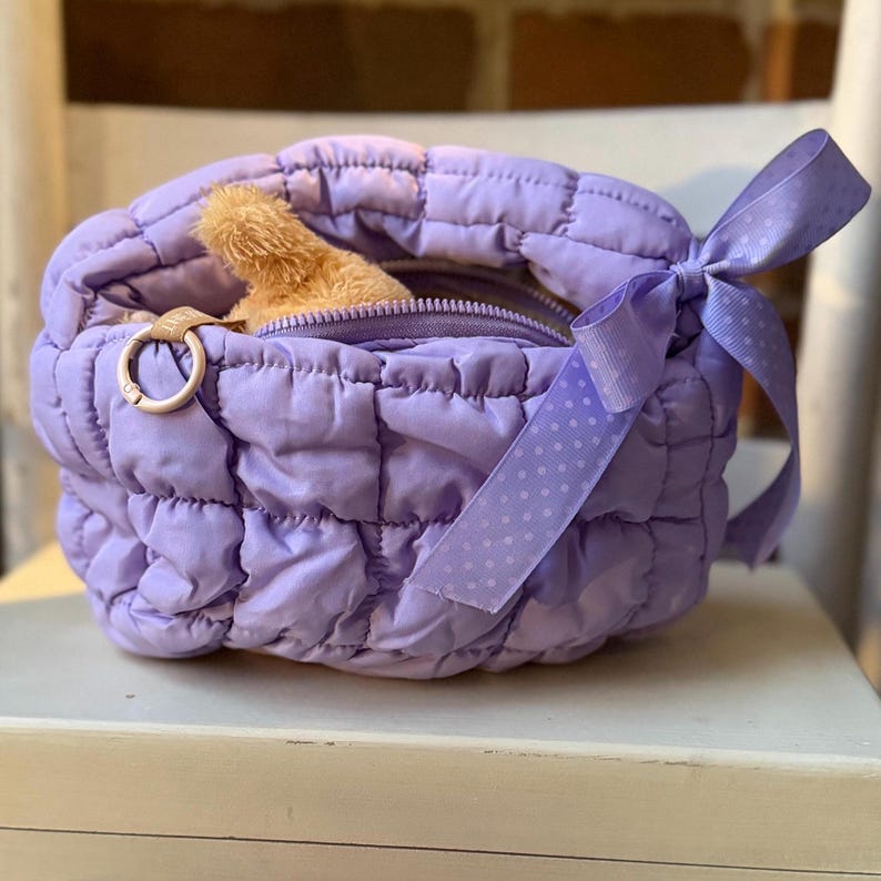May include: A quilted lavender bag with a zipper closure and a light brown stuffed animal peeking out. The bag has a matching lavender ribbon with white polka dots tied in a bow. A gold-colored ring is attached to the side.