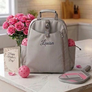 Op de afbeelding: Een grijze rugzak met de naam "Louisa" in script, een roze pickleball en een paddle. Een boeket roze rozen en een kaart met "Happy Mother's Day!" zijn ook aanwezig. De sc&egrave;ne speelt zich af op een wit aanrecht.