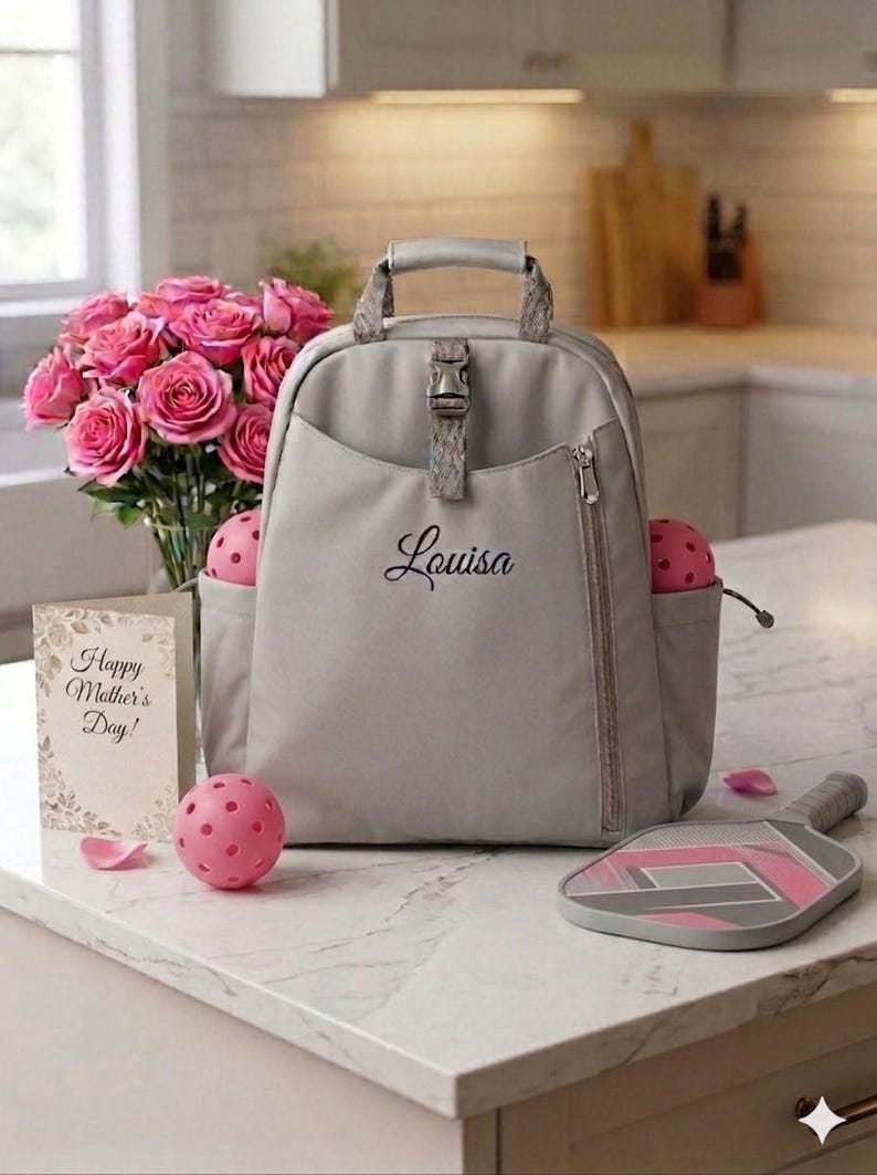 Op de afbeelding: Een grijze rugzak met de naam "Louisa" in script, een roze pickleball en een paddle. Een boeket roze rozen en een kaart met "Happy Mother's Day!" zijn ook aanwezig. De sc&egrave;ne speelt zich af op een wit aanrecht.