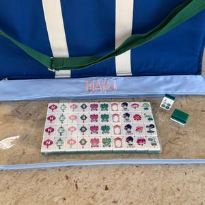 May include: A blue and white striped tote bag with a green strap and a light blue Mahjong game case. The case contains a set of tiles with various symbols and characters. Two dice and a green tile are also visible.
