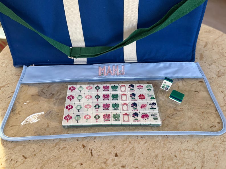 May include: A blue and white striped tote bag with a green strap and a light blue Mahjong game case. The case contains a set of tiles with various symbols and characters. Two dice and a green tile are also visible.