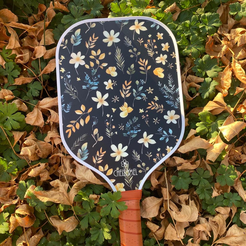 K&ouml;nnte beinhalten: Ein schwarz-wei&szlig;es Pickleball-Schl&auml;ger mit einem Blumenmuster. Der Schl&auml;ger hat einen wei&szlig;en Rand und der Text "CHELSEA MEL" ist auf dem Griff aufgedruckt.