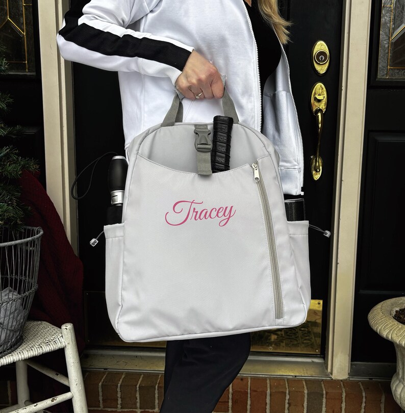 Op de afbeelding: Een lichtgrijze rugzak met een zijrits en zijvakken. Het woord "Tracey" is in roze script op de voorkant geborduurd. De rugzak heeft grijze banden en een zwarte gespsluiting.