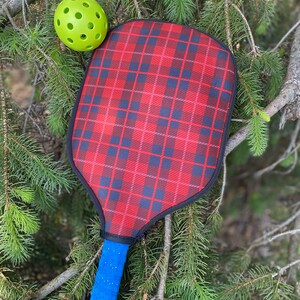 Pu&ograve; includere: Una racchetta da pickleball a scacchi rossi e neri con un manico blu e una palla da pickleball verde.