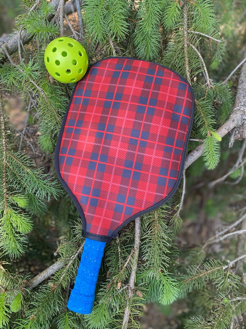 Pu&ograve; includere: Una racchetta da pickleball a scacchi rossi e neri con un manico blu e una palla da pickleball verde.