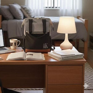 May include: A multi-compartment backpack in gray, black, and brown sits on a wooden desk, next to a laptop, a mug, and an open book. A white lamp with a beige lampshade and a stack of notebooks are also on the desk. The backpack has the initials "AK" on the front.
