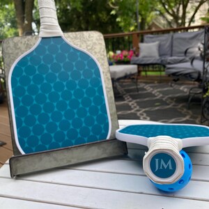 May include: Two teal blue pickleball paddles with white handles and a white ball with the initials "JM" on it. The paddles are on a white wooden table.
