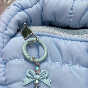 May include: Close-up of a light blue quilted fabric with a silver zipper and a light blue clasp. A light blue bow-shaped charm with a crystal bead hangs from the clasp. The charm is attached to a light blue ring.