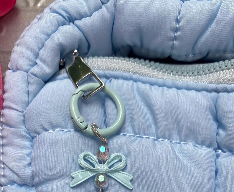 May include: Close-up of a light blue quilted fabric with a silver zipper and a light blue clasp. A light blue bow-shaped charm with a crystal bead hangs from the clasp. The charm is attached to a light blue ring.