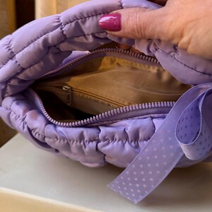 May include: A quilted, lavender-colored handbag with a zipper closure. The interior is a light tan color. A lavender ribbon with white polka dots is attached to the side of the bag.