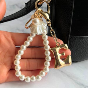 May include: A gold-coloured keyring with a large letter "E" charm. The keyring features a pearl beaded wristlet and a decorative pearl and shell accent. The keyring is attached to a black handbag.