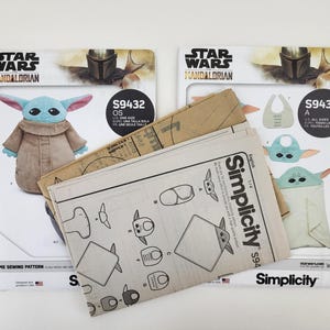 K&ouml;nnte beinhalten: Zwei Star Wars Mandalorian N&auml;hvorlagen. Ein Schnittmuster zeigt eine Grogu-Pl&uuml;schpuppe, das andere eine Grogu-L&auml;tzchen und eine Decke. Die Muster sind von Simplicity und enthalten den Text "S9432 OS" und "S9433 A".