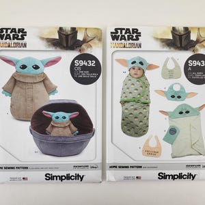 K&ouml;nnte beinhalten: Zwei Simplicity-Schnittmusterpackungen mit Star Wars Mandalorian-Designs. Eines zeigt eine Grogu-Pl&uuml;schpuppe und einen Tr&auml;ger, das andere Babyzubeh&ouml;r. Die Packungen enthalten den Text "STAR WARS MANDALORIAN" und "Simplicity".
