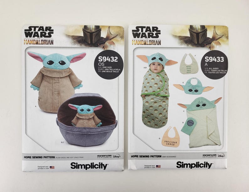 K&ouml;nnte beinhalten: Zwei Simplicity-Schnittmusterpackungen mit Star Wars Mandalorian-Designs. Eines zeigt eine Grogu-Pl&uuml;schpuppe und einen Tr&auml;ger, das andere Babyzubeh&ouml;r. Die Packungen enthalten den Text "STAR WARS MANDALORIAN" und "Simplicity".