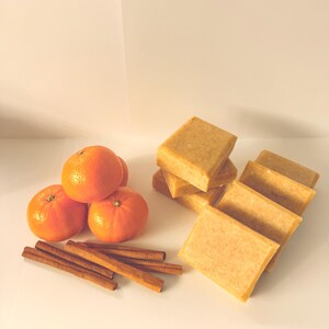 Puede incluir: Naturaleza muerta con naranjas, palitos de canela y pastillas de jabón rectangulares. Las naranjas son de color naranja brillante, los palitos de canela son marrones y los jabones son de color beige claro. Los artículos están dispuestos sobre una superficie blanca.