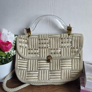 Może przedstawiać: Beżowa torebka w stylu vintage z tkanym wzorem i przezroczystą plastikową rączką. Torebka posiada złote zapięcie i wykończenia oraz pasujący pasek na ramię. Projekt to mieszanka beżu i złamanej bieli, tworząca teksturowany wygląd.