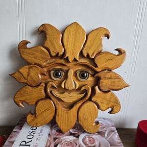 May include: Wooden sun face wall decoration with a smiling expression. The sun has a detailed carved design with petal-like rays and blue eyes. The piece is made of light brown wood and is displayed on a book with the word "Rose" on it.