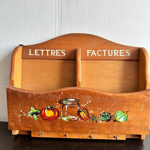 Peut inclure: Un organisateur mural en bois avec deux compartiments étiquetés "Lettres" et "Factures". L'organisateur a un motif peint de légumes et un bocal en bas et quatre crochets.
