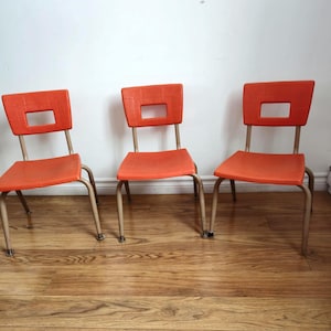 May include: Three orange plastic chairs with metal legs. The chairs have a rectangular back with a cut-out rectangle in the middle.