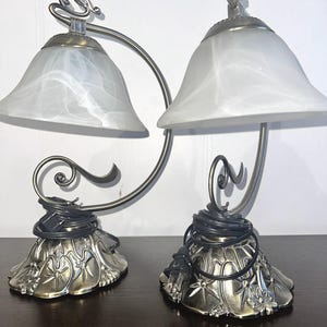 May include: A pair of antique-style table lamps with white alabaster shades and ornate metal bases. The lamps have a curved metal arm connecting the two shades.