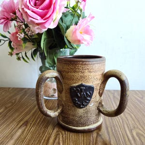 Puede incluir: Una taza de cerámica marrón con dos asas y un escudo marrón y negro con el texto "Prov. of Isle" en él.