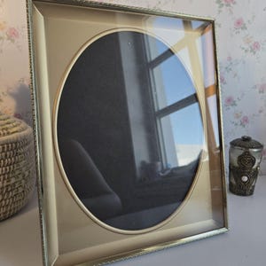 Peut inclure: Un cadre photo carré avec une finition dorée métallique et un bord extérieur texturé et perlé. Le cadre présente une grande ouverture ovale centrée avec un passe-partout de couleur sombre à l'intérieur, protégé par une vitre transparente.