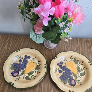 May include: Two yellow and green ceramic plates with a design of grapes, plums, and pears. The plates have a scalloped edge and a green rim.