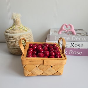 Peut inclure: Un panier carré tressé rempli de nombreux objets ronds rouges, peut-être des cerises. Le panier est de couleur marron clair et possède deux poignées arquées. Un récipient décoratif tissé et des livres sont en arrière-plan.