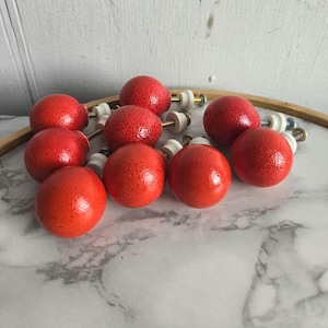 Peut inclure: Dix boutons ronds en bois rouge avec des bases en plastique blanc et des vis dorées. Les boutons sont disposés en grappe sur une surface en marbre blanc.