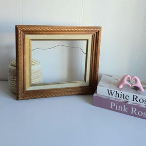 Peut inclure: Un cadre photo rectangulaire vide avec une bordure dorée ornée. Un panier en osier et deux livres, "White Rose" et "Pink Rose", sont à côté du cadre. Une sculpture de mains roses en forme de cœur est posée sur les livres.