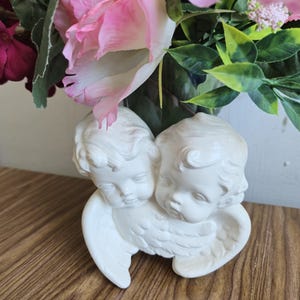 Könnte beinhalten: Eine weiße Cherub-Figur mit zwei cherubischen Gesichtern und gefiederten Flügeln. Die Figur steht auf einer Holzoberfläche, mit einem Hintergrund aus rosa und roten Blumen und grünen Blättern.