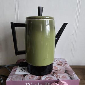 Puede incluir: Una cafetera eléctrica verde vintage con asa, boquilla y base negras. La cafetera está sobre un libro con rosas rosas y las palabras "Pink Roses". Una estatua de mano rosa está en primer plano.