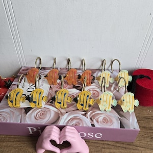 May include: A collection of shower curtain hooks with tropical fish designs. The hooks are in shades of yellow, orange, and white, with black and blue details. The hooks are displayed on a box with a floral pattern.