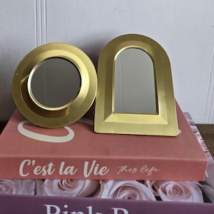 Peut inclure: Deux miroirs décoratifs dorés, un rond et un en forme d'arche, reposent sur une pile de livres. Le miroir rond a un cadre circulaire, tandis que le miroir en forme d'arche a une base rectangulaire. Les livres sont roses et violets avec le texte "C'est la Vie" visible.